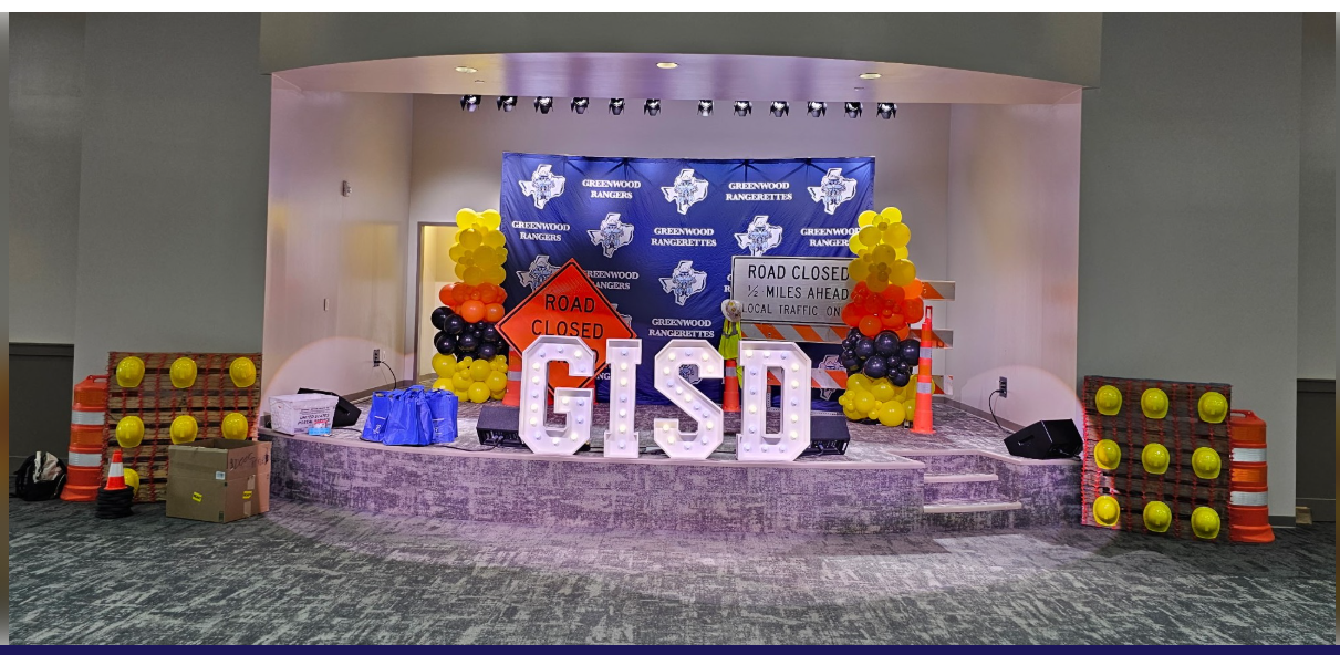 Greenwood ISD event stage with large GISD marquee letters, a Rangers and Rangerettes backdrop, and a construction theme of road-closed signs, hard hats, and traffic cones.